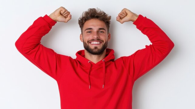 A young man with a beard and curly hair is smiling broadly and flexing his biceps. He is wearing a red hooded sweatshirt