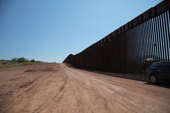View of the steel bollard border wall stretching along a dirt road under a clear blue sky in Naco, Arizona, United States.