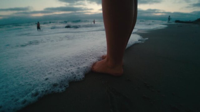 Intimate low-angle close-up of bare feet planted in wet sand as sea foam swirls around the ankles at dusk, distant beachgoers, mindful coastal moment, summer seaside calm.