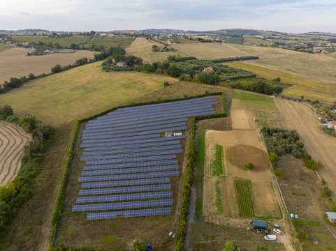 Aerial view of a solar farm with rows of photovoltaic panels surrounded by green hedges and agricultural fields in the rural landscape of Fermo, Marche, Italy.