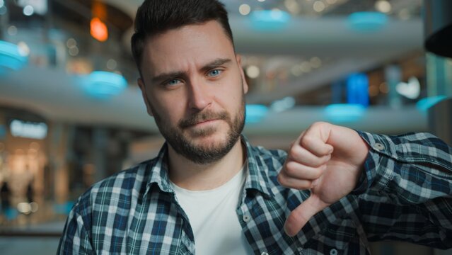 Unhappy Caucasian man showing thumb down gesture looking at camera with negative opinion disagreement bad news in shopping mall business male client sad businessman demonstrate dislike sign indoors