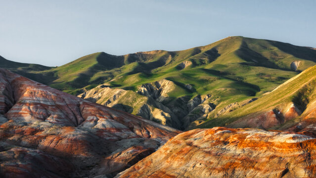 View of the colorful striped landscape of the Candy Cane Mountains with rolling green hills and red clay formations under a clear sky in Azerbaijan.