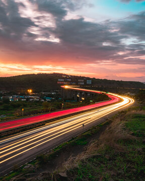 View of the Mumbai-Pune Expressway at sunset with vibrant light trails from moving vehicles, surrounded by rolling hills and a dramatic cloudy sky in Pune, Maharashtra, India.