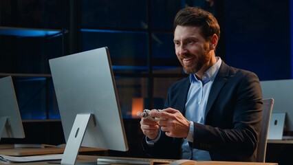 Funny Caucasian middle-aged business man computer work pause playing online game at night evening dark office male businessman looking around taking joystick play console winning optimistic relax © Yuliia