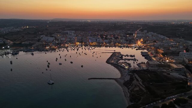 Marsaxlokk, Malta fishing village festa with illuminated harbor, anchored boats and parish church at dusk along the Mediterranean waterfront, captured by drone.