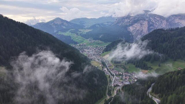 Aerial drone view overlooking Selva di Val Gardena at the beginning of Gardena Pass, with the mountain town surrounded by forested slopes, green valleys, rugged peaks, and drifting clouds