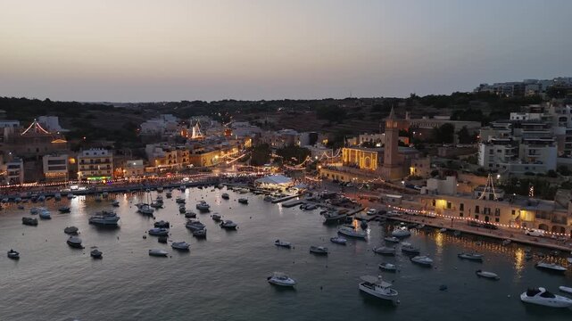 Marsaskala Bay, Malta parish church and marina with docked boats, waterfront apartments and illuminated village festa streets at dusk, captured by drone.