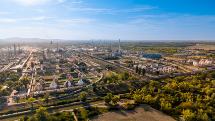 Obraz premium Aerial view of an oil refinery with spherical storage tanks and steel pipelines. Industrial complex features gas spheres, tall chimneys, green trees, bright sunlight, clear blue sky.