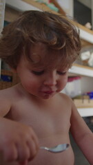 Fototapeta premium Toddler eating independently with spoon while sitting on kitchen floor, candid childhood mealtime highlighting self feeding skills, curiosity, confidence and healthy development