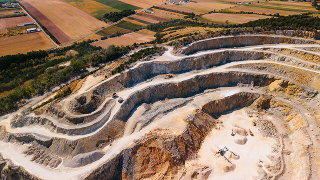 Terraced sand quarry pit with excavators working on ledges. Stepped rock walls surrounding deep pit with heavy machinery, dirt paths and fields.