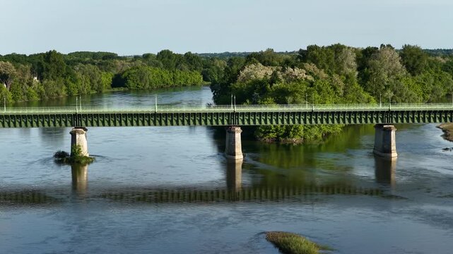 The Pont Canal bridge spanning across a river in Briare, France. The bridge is supported by stone pillars and surrounded by lush green trees and foliage on both sides of the river, wide shot.