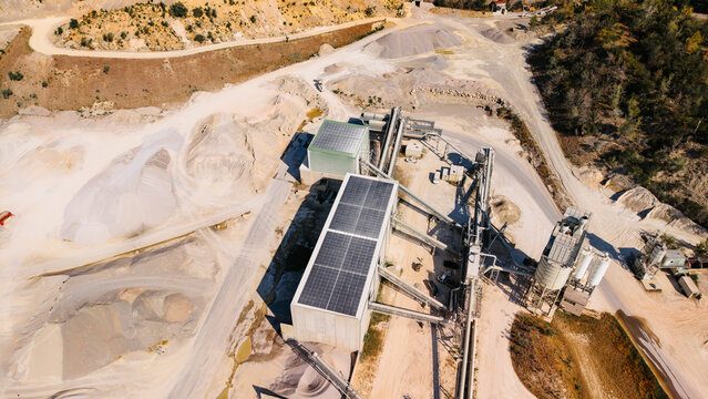 Aerial view of sand quarry processing facility with solar panels. Modern aggregate plant with rooftop solar panels silos conveyor systems and surrounding terrain.