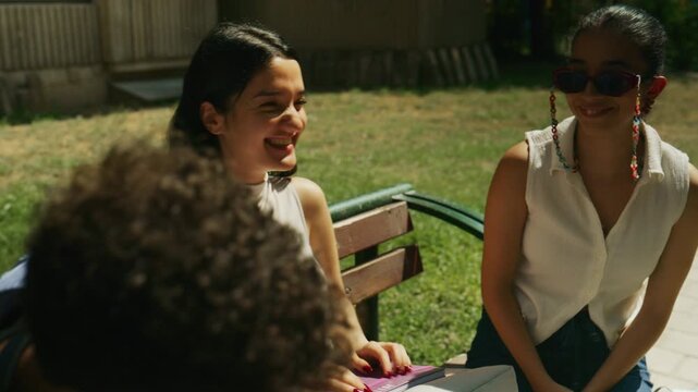 High school students are sitting on a bench outdoors, smiling and laughing together. Young friends share a moment of joy and conversation in a bright outdoor setting.