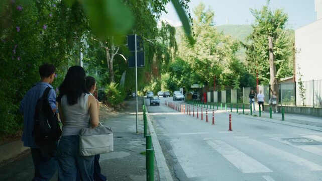 High school students stand on a sidewalk, waiting to cross a busy street. Three young friends are ready for their day on a vibrant campus.