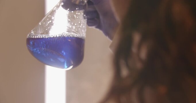 Over-the-shoulder video of a person shaking an Erlenmeyer flask filled with blue liquid in a laboratory setting. Scientific chemistry, experimentation, research, analysis