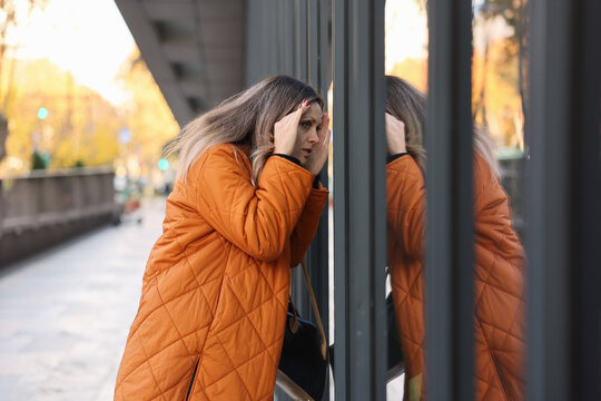 A woman in an orange coat peering through metal bars on an urban sidewalk, evoking a sense of curiosity and movement in the cityscape. The scene captures the essence of urban life with its.