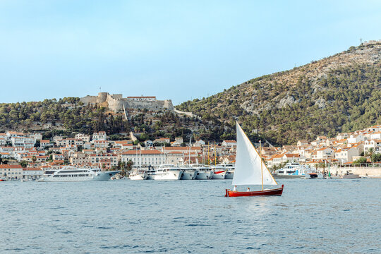View of Hvar Port with a small red sailboat sailing in the foreground, featuring historic buildings and the Spanjola Fortress on the hill in Hvar, Split-Dalmatia County, Croatia.