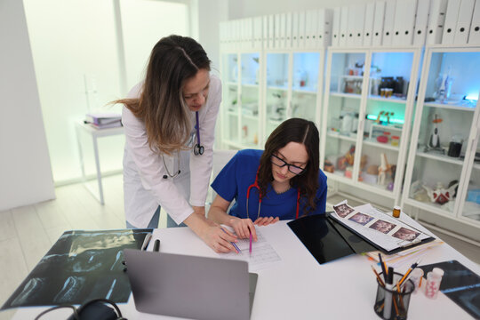 Female doctor in white coat and nurse in blue scrubs lean over desk, reviewing patient charts, ultrasound images, and heart rhythm printout inside bright modern clinic, showing teamwork, clinical