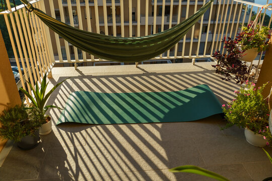 Sunlit apartment balcony featuring a green hammock, turquoise yoga mat, potted plants and strong geometric shadows cast by the railing.