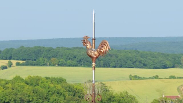 A traditional metallic rooster weathercock mounted atop a church spire, set against a backdrop of rolling green fields and dense forest, close-up shot.