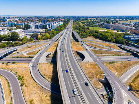 Roads intersect near a city in bright sunlight during the day with buildings and trees visible in the background
