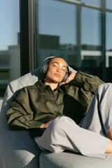 Fototapeta premium Person relaxing with headphones reclined on a gray lounge seat in bright sunlit interior for meditation and relaxation background and lifestyle banner