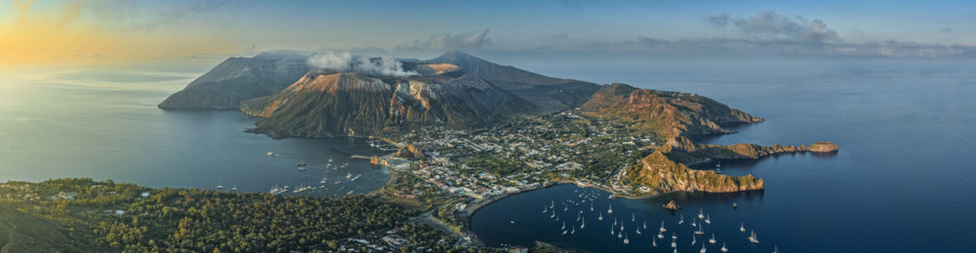 Aerial view of the volcanic island of Vulcano with its smoking crater, coastal town, and numerous boats anchored in the bay at golden hour in Lipari, Sicily, Italy.