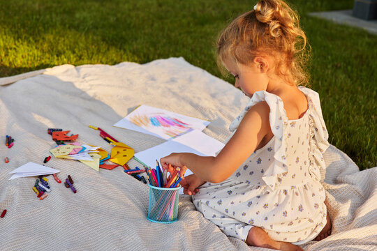 A young child sits on a blanket surrounded by crayons and paper. She focuses on her drawing while the sun shines in the background during late afternoon