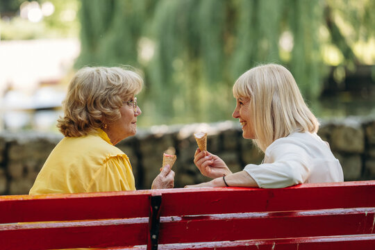 Two senior women enjoying ice cream cones while sitting on a red park bench surrounded by greenery and a serene water view in a public park setting