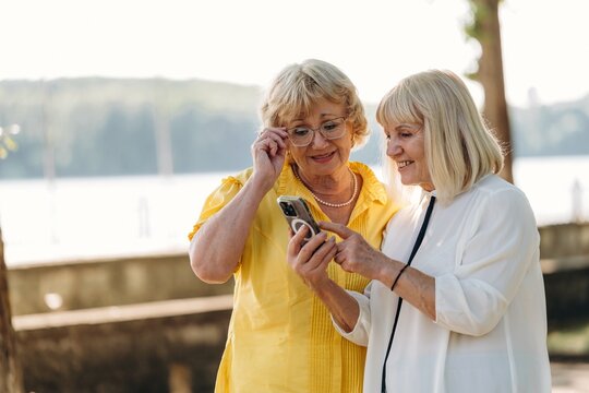 Two senior women with glasses and light-colored hair share a moment while looking at a smartphone outdoors by a lake with trees in the background