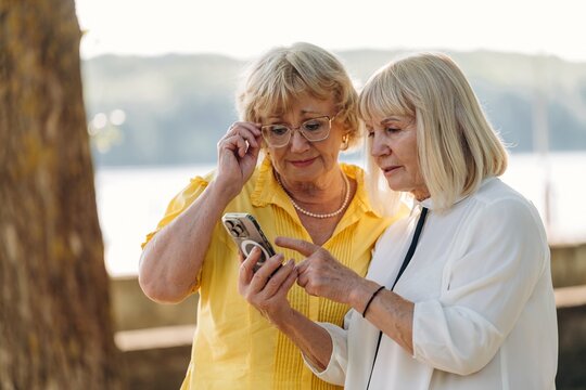 Two senior women with glasses sharing a smartphone outdoors, one wearing a yellow blouse and the other in a white top, with a natural background of trees and water