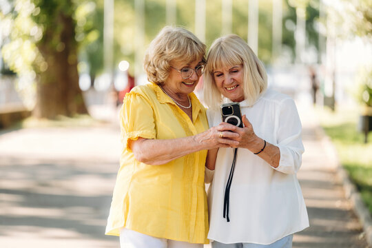 Two senior women sharing a moment while looking at a camera in a sunny park with trees and a pathway in the background