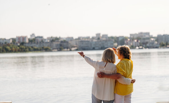 Two women standing by a lake, one pointing towards the city skyline while the other embraces her, showcasing a serene waterfront view with buildings in the background