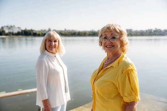Two senior women smiling by the lakeside, one in a white blouse and the other in a yellow shirt, with calm water and cityscape in the background