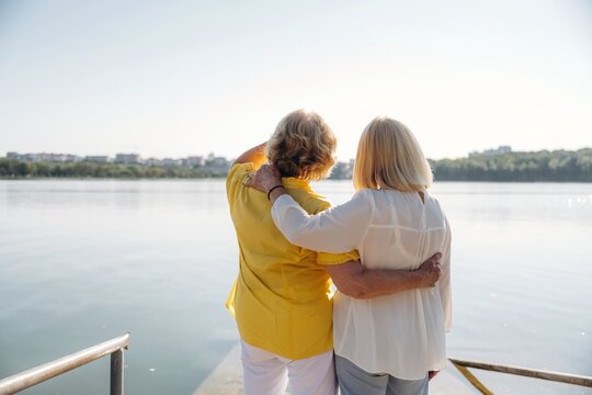 Two senior women embracing each other while standing on a pier overlooking a calm lake with a clear sky and distant cityscape in the background
