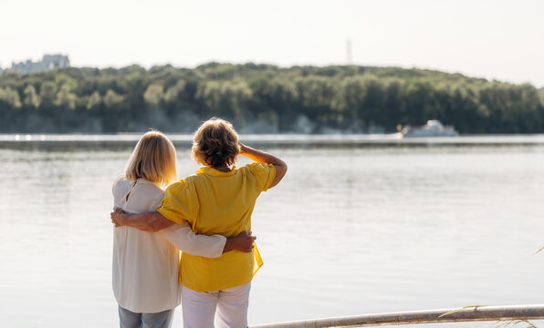 Two women standing by a lake, one in a yellow shirt and the other in a white blouse, enjoying the view of the water and trees on a sunny day