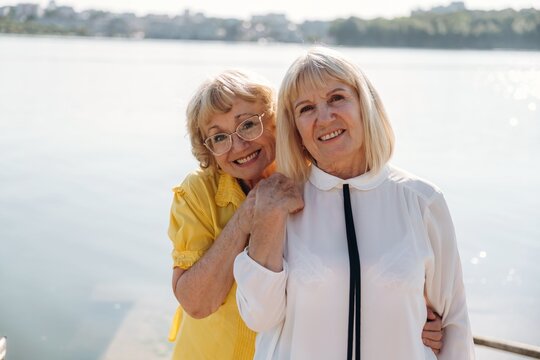 Two senior women smiling together by a lake, one wearing yellow shirt and glasses, the other in white blouse, with water and city skyline in the background