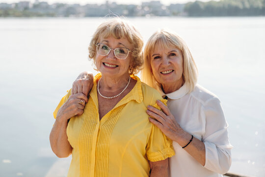 Two smiling senior women standing together by a lake, one wearing a yellow blouse and the other in a white top, with a serene water background and soft sunlight