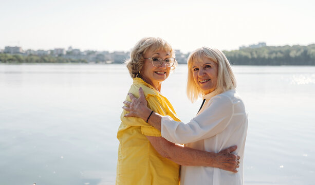 Two senior women smiling and embracing each other by the lakeside, wearing colorful outfits with a serene water backdrop and city skyline in the distance