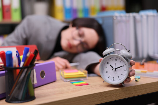 Tired businesswoman asleep at desk with white alarm clock, pens, notes, folders, and sticky tabs in blurred office background, suggesting burnout, time pressure, study fatigue, and missed rest