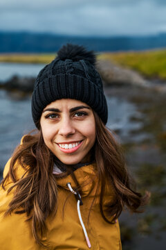 A white woman smiles outdoors in cold weather near a rocky shore