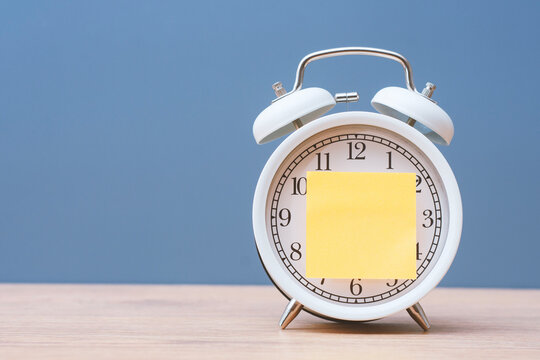 White alarm clock with a blank yellow sticky note on a wooden table, representing time management, reminders, deadlines, scheduling, productivity and daily planning.