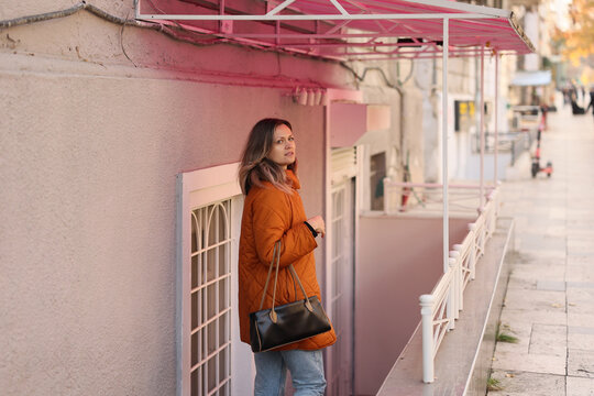 Young woman pauses beside pastel stucco facade, wearing orange quilted coat and holding black handbag under pink awning along quiet urban sidewalk with soft daylight, calm winter mood, shallow depth