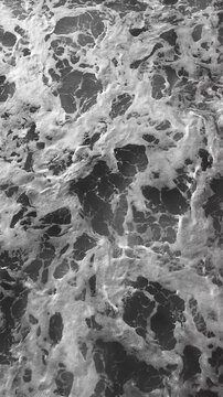 Black and white close-up of powerful ocean waves with swirling sea foam creating abstract natural patterns and dynamic textures on moving water surface. Vertical video
