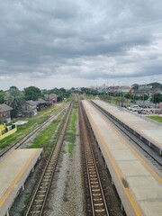 Obraz premium A high-angle elevated view showing long parallel railway tracks stretching through an empty train station platform with a cloudy sky and rural landscape background.