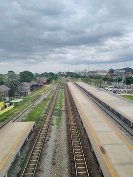 A high-angle elevated view showing long parallel railway tracks stretching through an empty train station platform with a cloudy sky and rural landscape background.