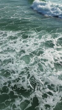 Aerial top view of turquoise ocean waves with white sea foam creating dynamic natural patterns and textures on moving seawater surface during a bright sunny summer day. Vertical video