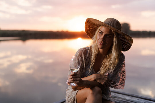 Woman Enjoys a Drink by the River During Summer at Sunset With a Large Hat