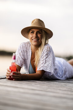 Woman Enjoys a Drink by the River During Summer With a Straw Hat on a Sunny Day While Lying on a Dock