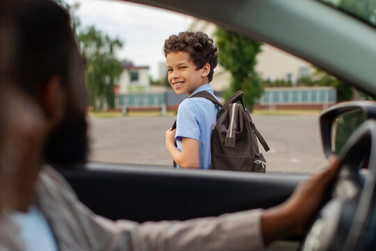Black schoolboy with backpack looking back at father driving him to school